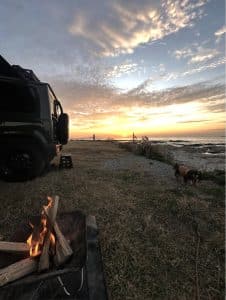 夕暮れの海辺、ジムニーの横で焚き火。犬が歩いている